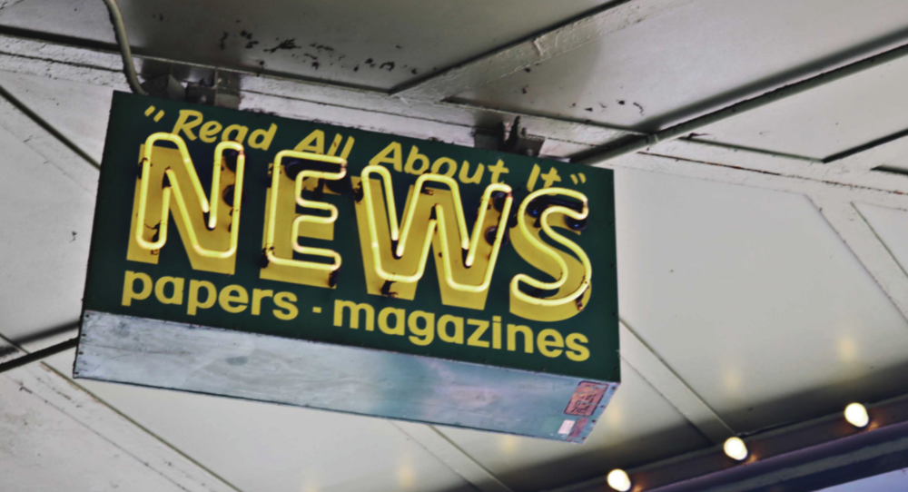 neon sign hanging from ceiling that reads "read all about it: news papers and magazines"